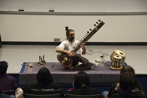 man playing sitar on blanket with drums and gold artifacts on lecture hall stage